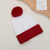 A festive knitted beanie in white with a red pom-pom and a matching red cuff, displayed next to a wooden surface.