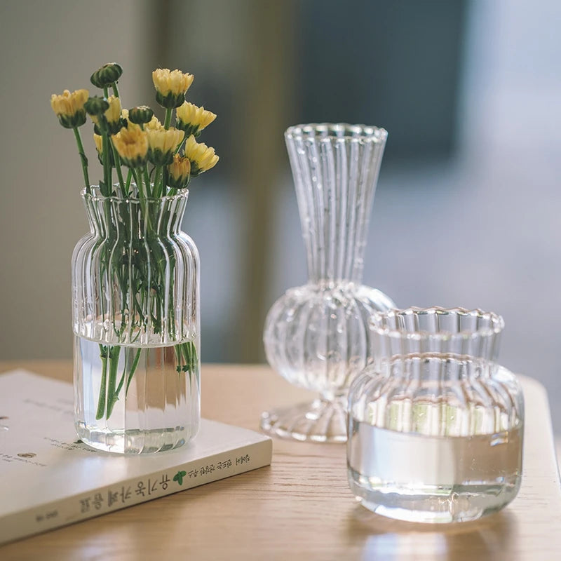 A clear glass vase filled with yellow flowers sits next to a vintage striped glass decanter and a book on a wooden table.