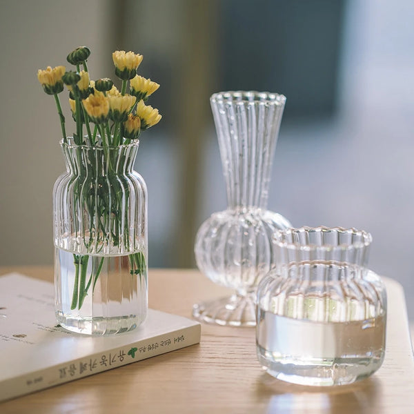 A clear glass vase filled with yellow flowers sits next to a vintage striped glass decanter and a book on a wooden table.
