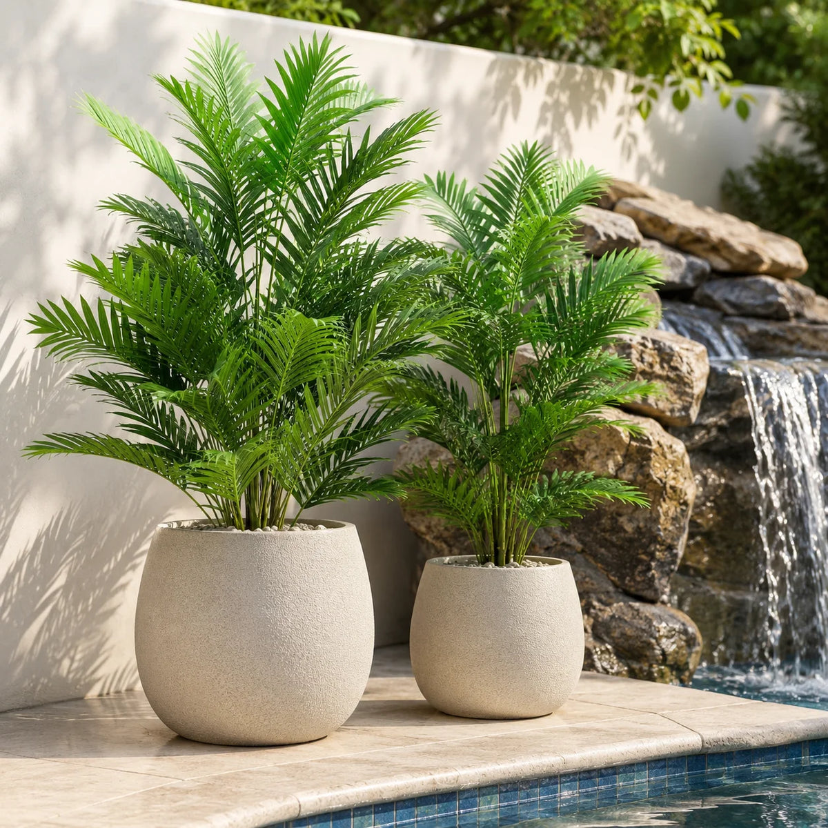 Two artificial areca palm trees in light gray pots sit on a stone ledge next to a water feature.