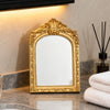 A vintage arched carved gold vanity mirror with a reflective surface displayed on a marble countertop.