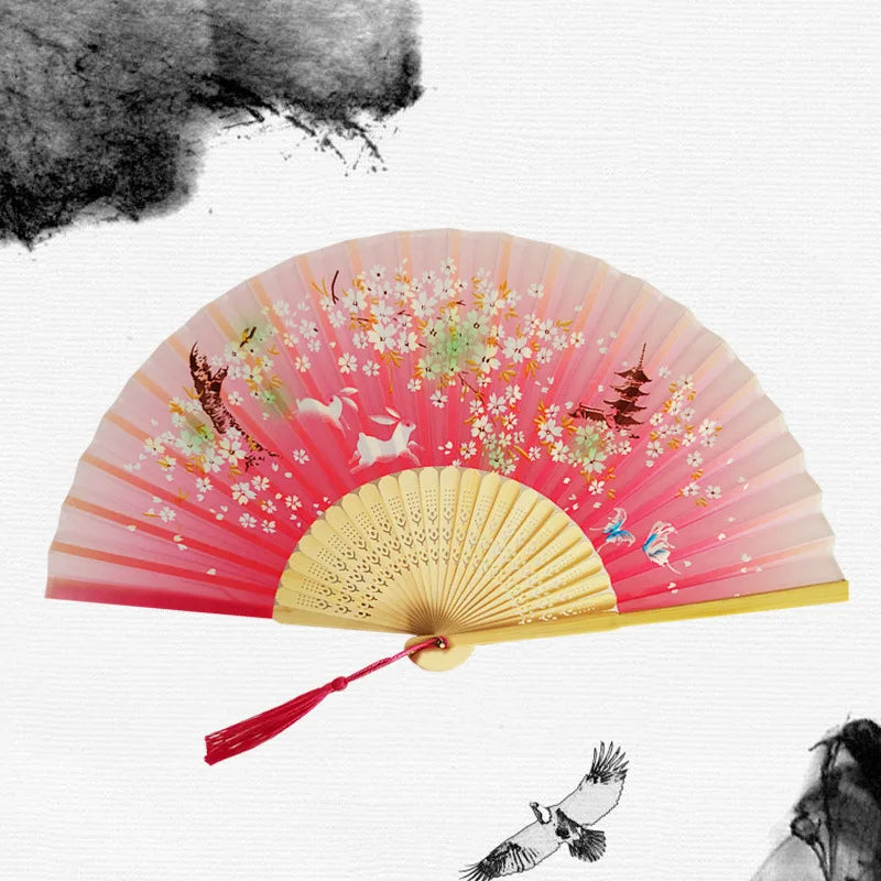 A vintage pink and gold bamboo folding hand fan with floral accents against a white background.