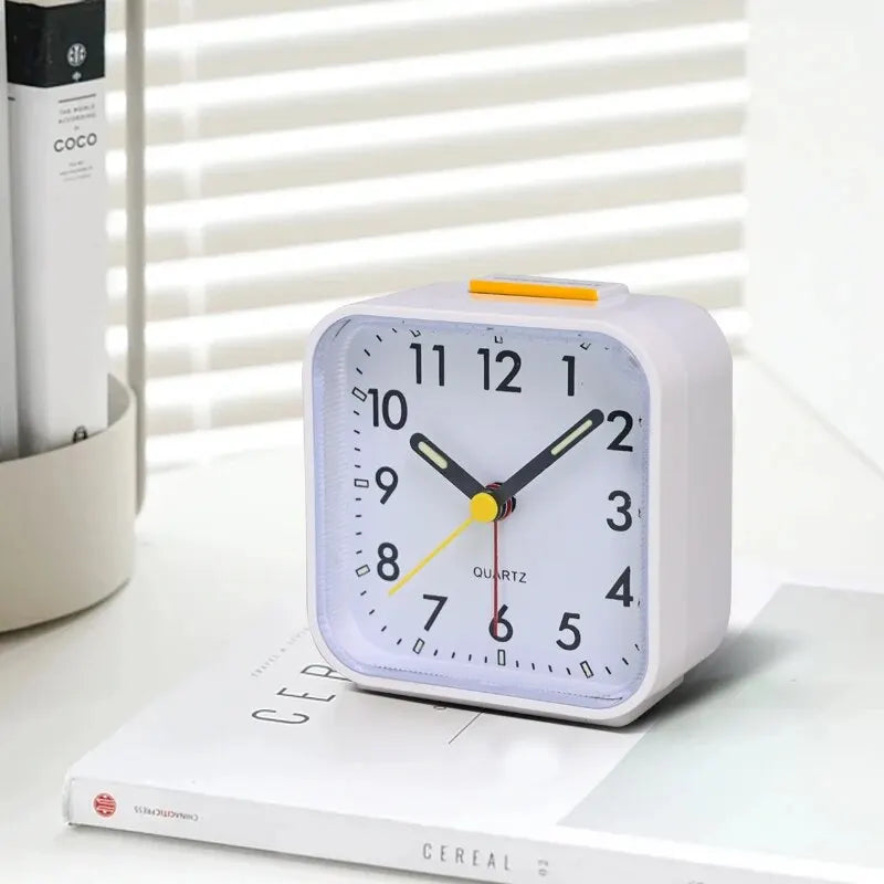 A white digital alarm clock with a light-colored face and orange hands is displayed on a white surface.