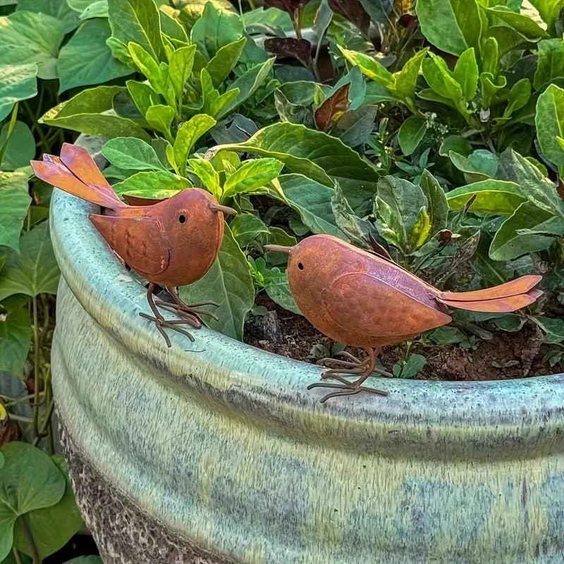 A small brown bird sculpture sits in a green ceramic pot surrounded by lush green foliage.