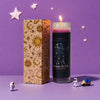 A glass candle in a cylindrical jar with a dark blue wax and a gold label featuring zodiac symbols, set against a purple back