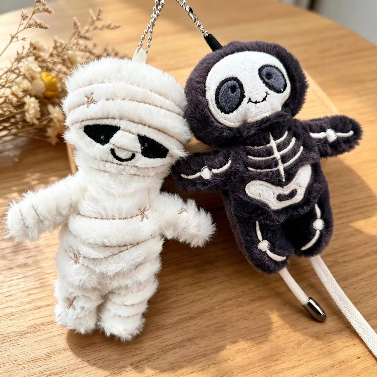 A white mummy skeleton plush keychain next to a dark gray skeleton plush keychain on a wooden surface.