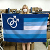 A person holds up a dark blue and light blue Pride flag banner with white symbols and stripes in a warehouse setting.