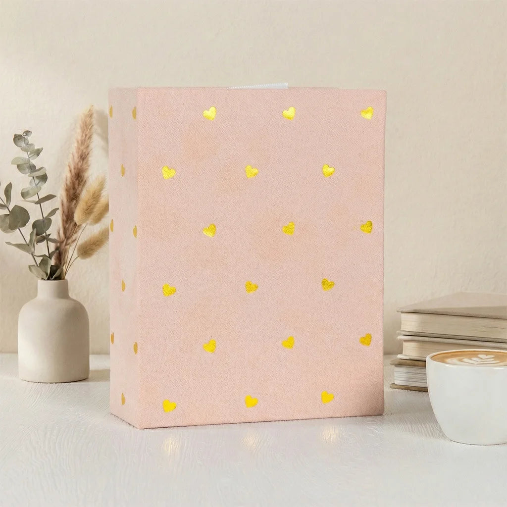 A pink photo album with gold foil hearts rests on a white surface next to a vase of dried flowers.