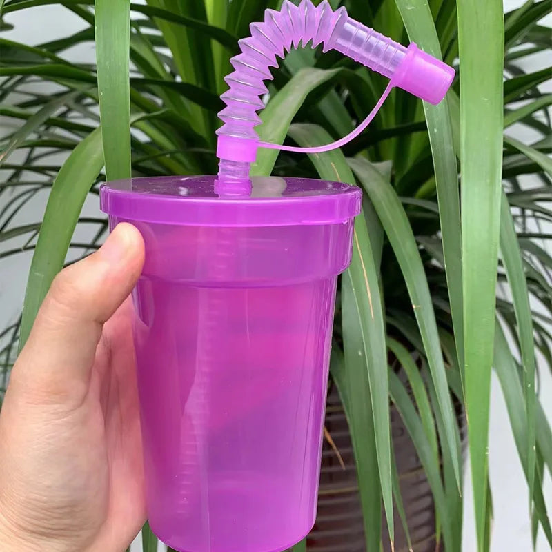 A hand holds a purple reusable sipper cup with a clear straw and lid, set against a background of green foliage.
