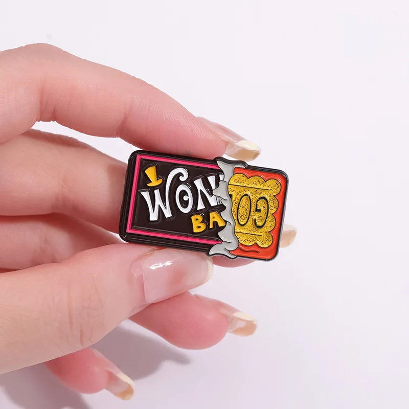 A hand holds a black enamel lapel pin featuring gold lettering and an orange border.