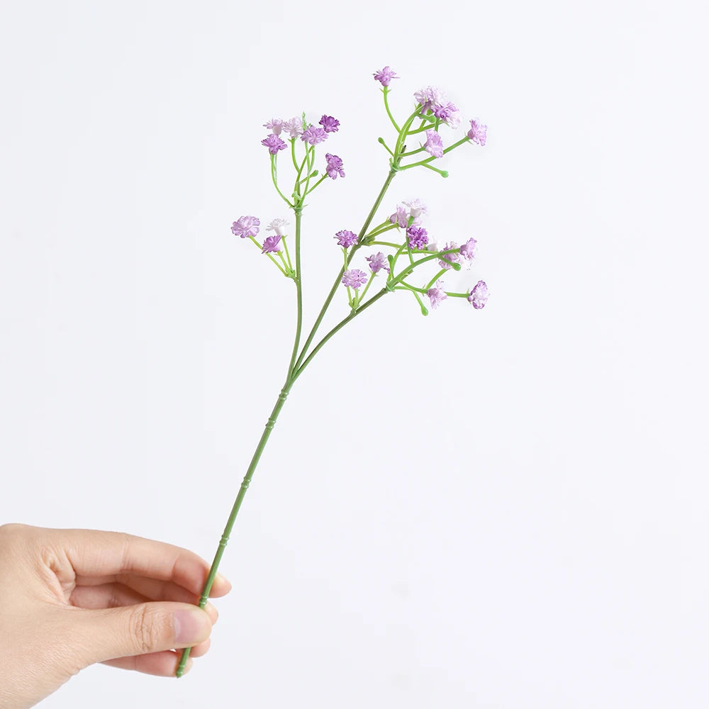 A hand holds a sprig of artificial baby's breath flowers with delicate purple florets.