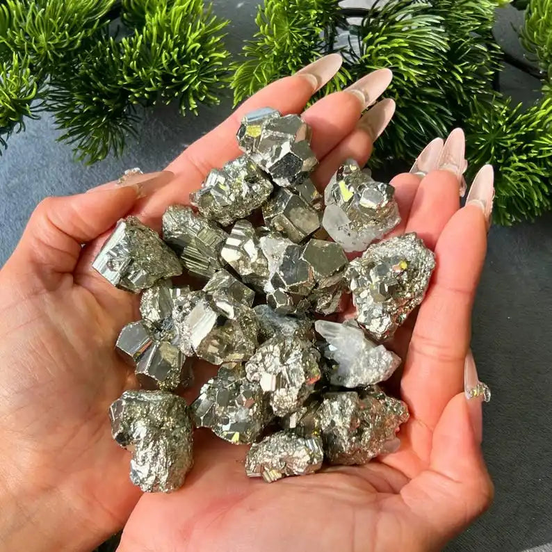 A hand holds a cluster of sparkling natural pyrite crystals against a backdrop of green foliage.