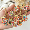 A hand holds a gold-plated charm bracelet featuring small religious icons set in colorful beadwork.