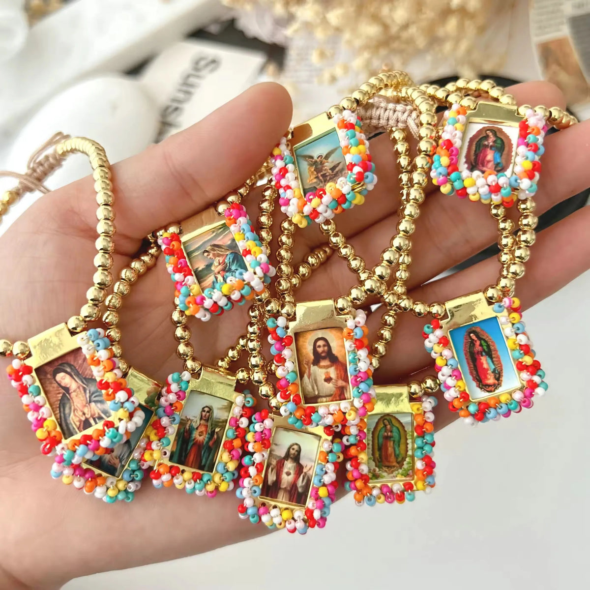 A hand holds a gold-plated charm bracelet featuring small religious icons set in colorful beadwork.