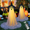 A glowing white ghost decoration with a black hat and sheer fabric is displayed outdoors in a garden setting.