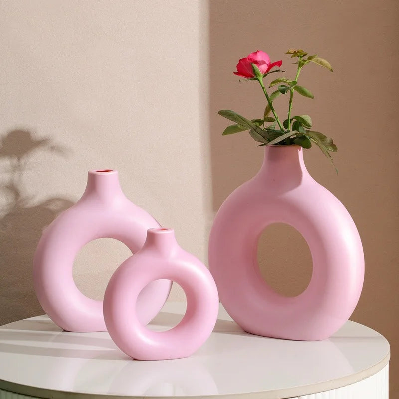 A pink ceramic vase with a circular opening holds a single red rose on a white surface.