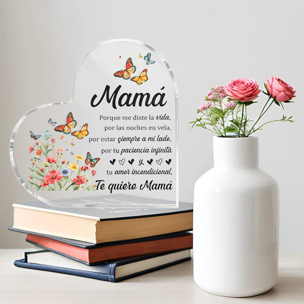 A clear heart-shaped ornament with a floral design and text in Spanish about love for mom sits next to a white vase and a sta