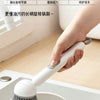 A hand is using an adjustable long-handled liquid washing brush to clean a frying pan with soap in a kitchen setting.