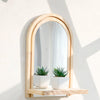 A wooden arched mirror framed in light wood mounted on a white wall with small potted plants.
