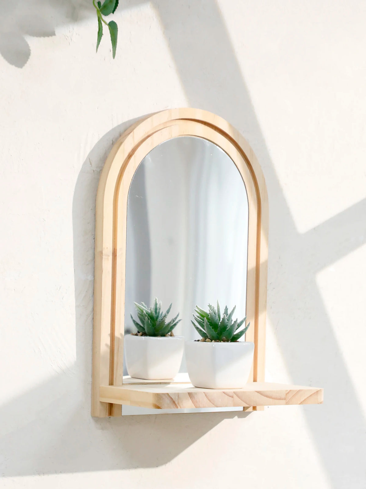 A wooden arched mirror framed in light wood mounted on a white wall with small potted plants.
