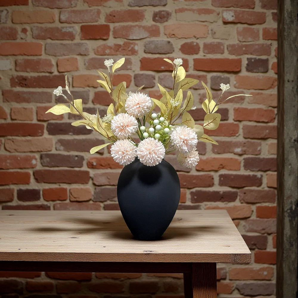 A dark vase holds a bouquet of light pink and cream fluffy flowers against a rustic brick background.