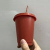A hand holds a reddish-orange plastic cold cup with a lid and straw against a light gray wall.