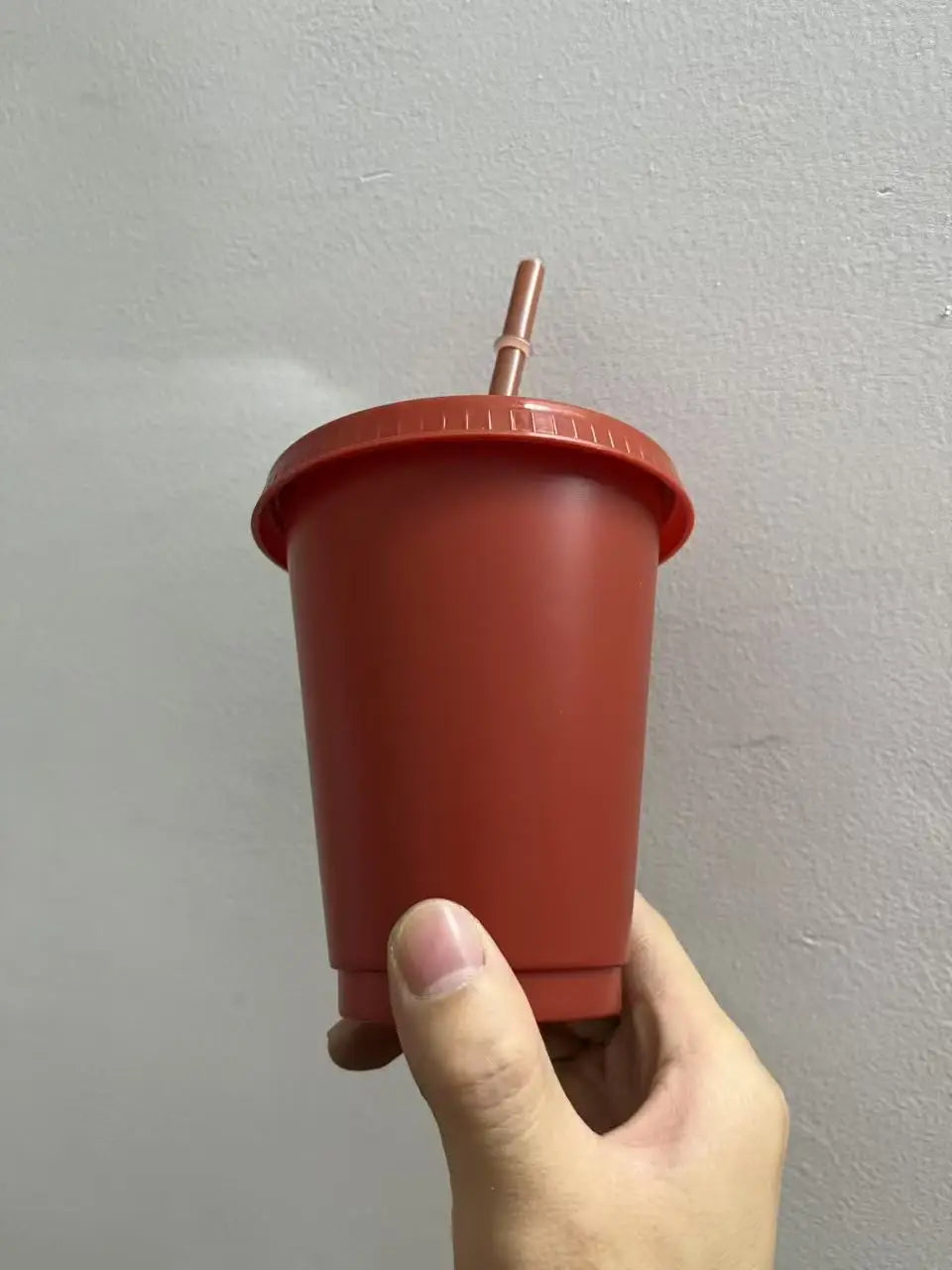 A hand holds a reddish-orange plastic cold cup with a lid and straw against a light gray wall.