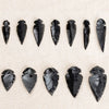 A collection of natural obsidian arrow pendants in various sizes and shapes arranged on a light fabric background.