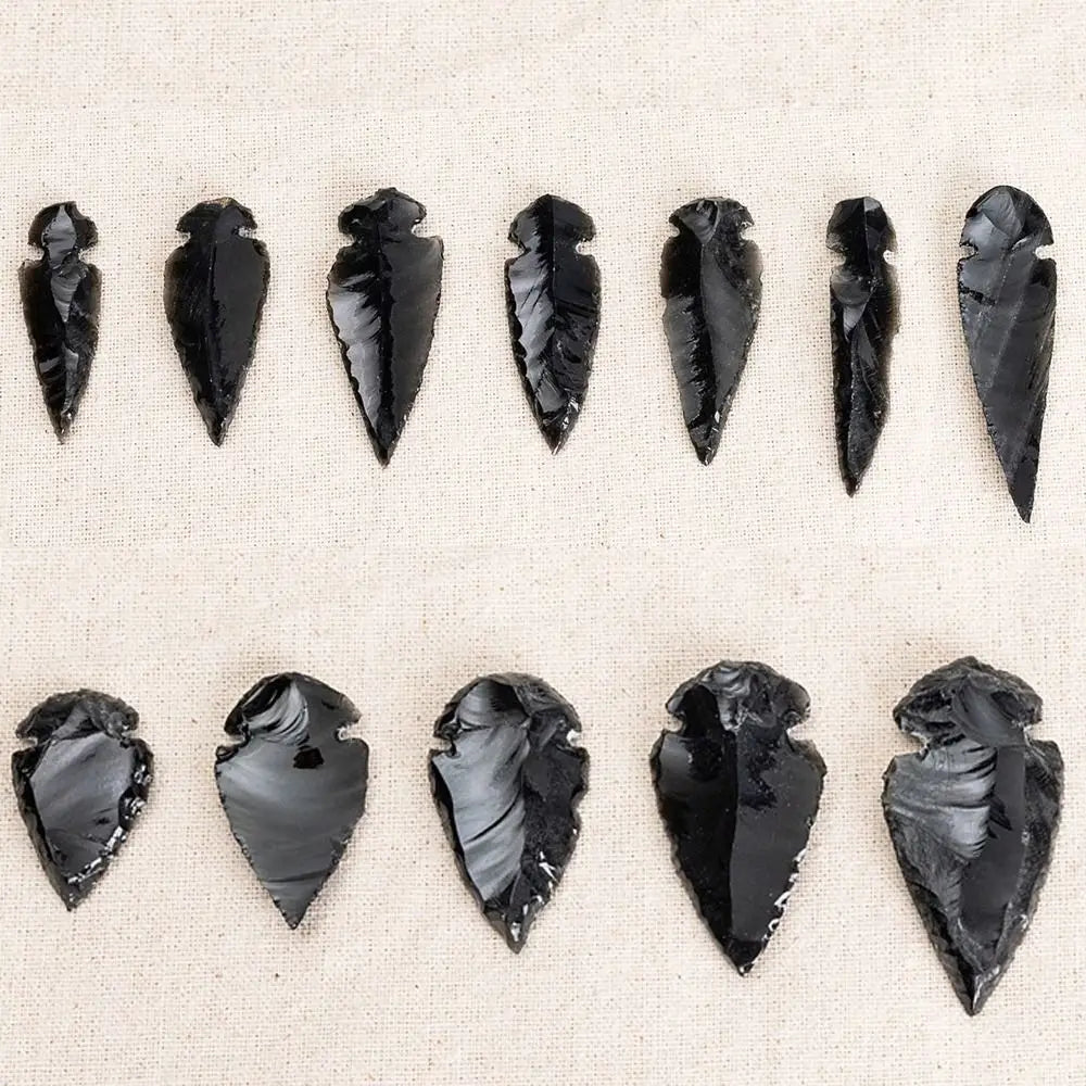 A collection of natural obsidian arrow pendants in various sizes and shapes arranged on a light fabric background.