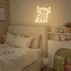 A bedroom scene featuring a white bed and a glowing neon sign shaped like a Highland cow on the wall.