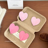 A set of pink heart-shaped adhesive key hooks displayed in a cardboard tray on a wooden surface.