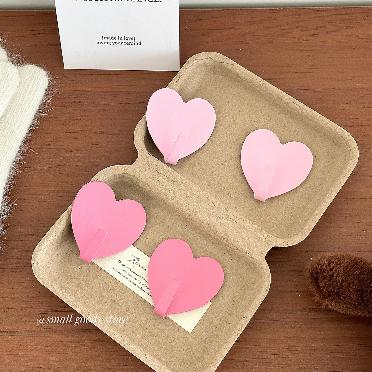 A set of pink heart-shaped adhesive key hooks displayed in a cardboard tray on a wooden surface.
