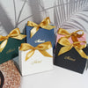 A collection of elegant paperboard thank you gift bags in various shades of blue, green, and black tied with gold ribbon.