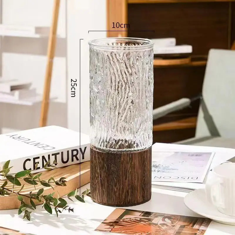 A clear glass vase with a textured surface sits on a dark wooden base, displayed on a white desk.