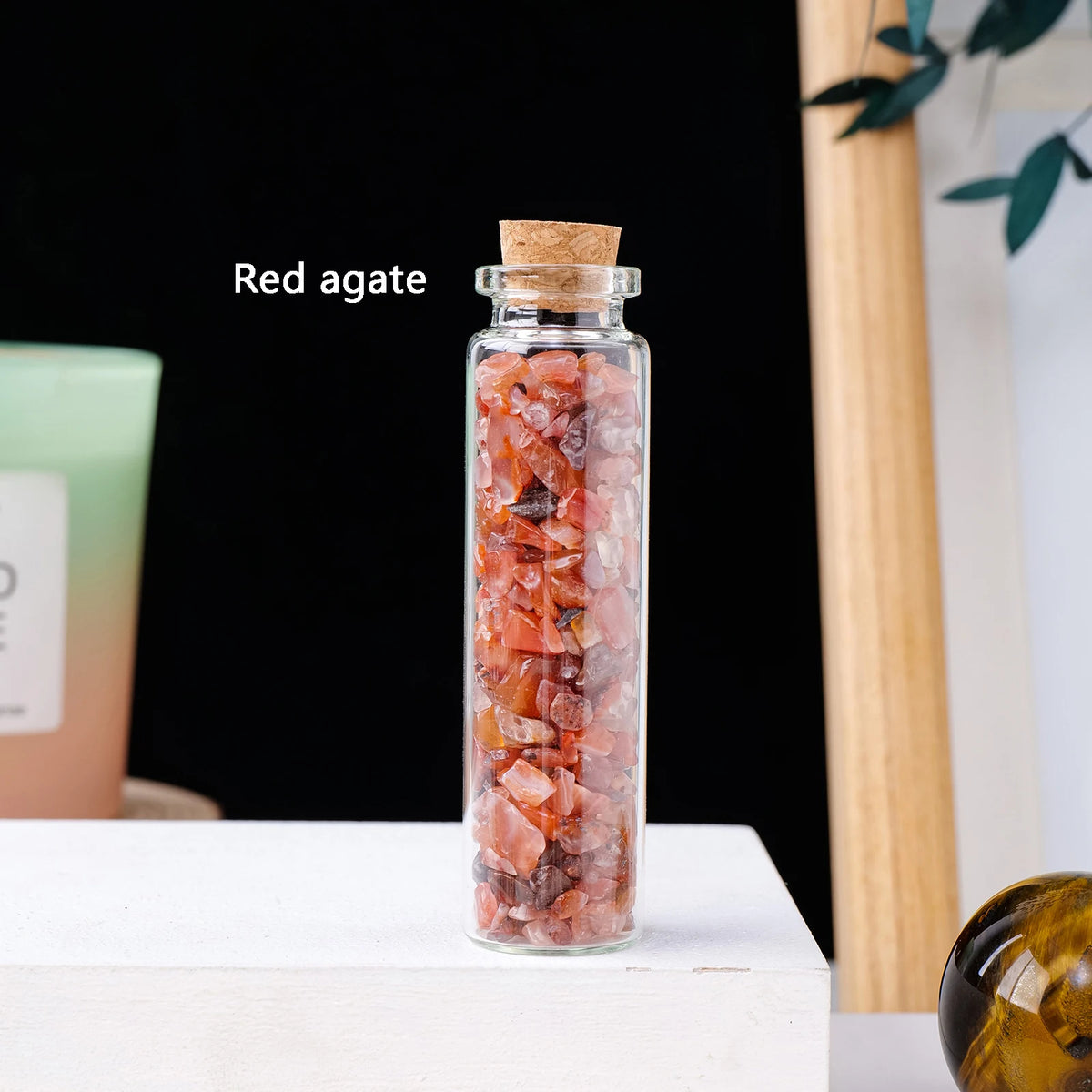 A glass bottle filled with red agate crystals sits on a white surface against a dark background.