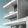 A modern, minimalist concrete building with stacked balconies and glass railings against a clear sky.