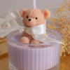 A soft pink teddy bear candle in a glass jar with a white ribbon, set against a blurred background of yellow flowers.