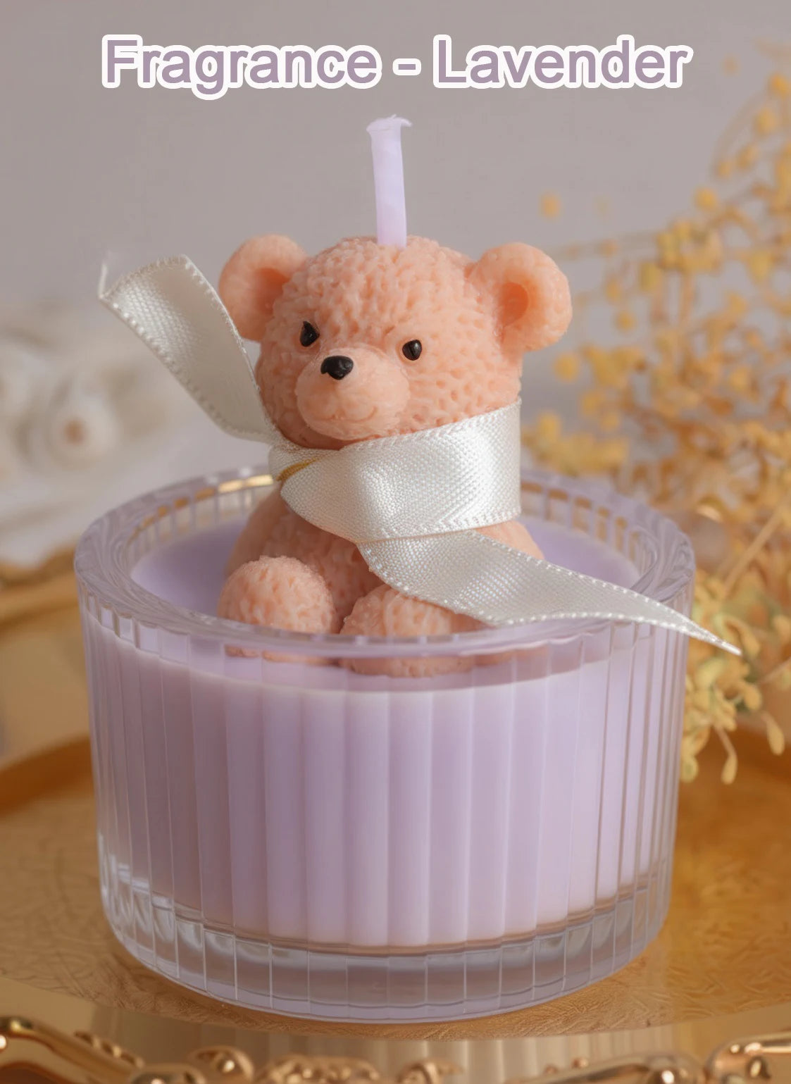 A soft pink teddy bear candle in a glass jar with a white ribbon, set against a blurred background of yellow flowers.