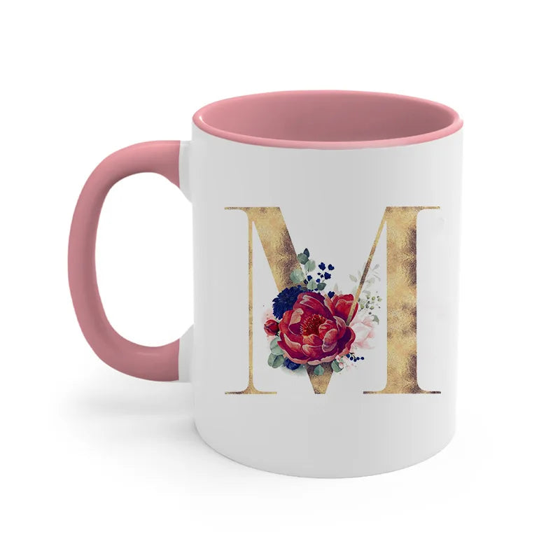 A pink ceramic coffee mug featuring a large gold letter M decorated with floral accents.