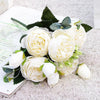 A bouquet of white silk rose and peony artificial flowers with green leaves on a light surface.