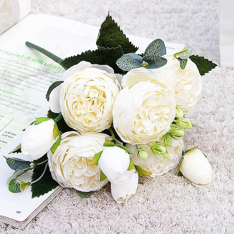 A bouquet of white silk rose and peony artificial flowers with green leaves on a light surface.