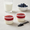 Three clear disposable dessert cups with lids containing layered red and white desserts next to a glass of milk and blueberri