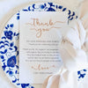 A thank you card with elegant script and floral blue patterns is placed on a white plate surrounded by white fabric.