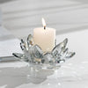 A white pillar candle with a flame sits in a clear crystal lotus-shaped holder on a white surface.