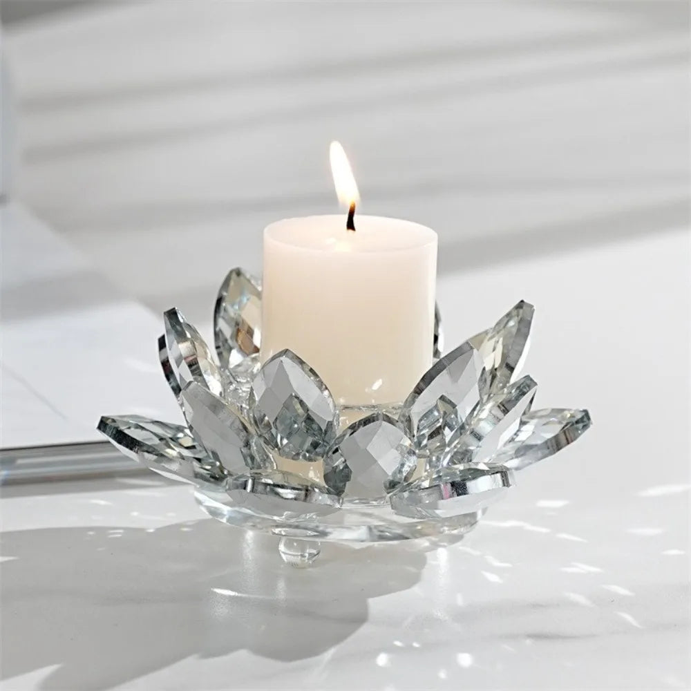 A white pillar candle with a flame sits in a clear crystal lotus-shaped holder on a white surface.