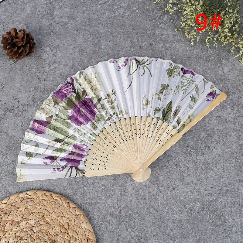 A hand carved bamboo folding fan with a floral and greenery pattern is displayed on a gray surface.