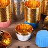 A collection of shiny gold greaseproof cupcake liners in various sizes and colors are displayed on a wooden surface.