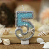 Glitter number cake topper shaped like the number 5 with a starfish design, set against a blurred background of seashells.