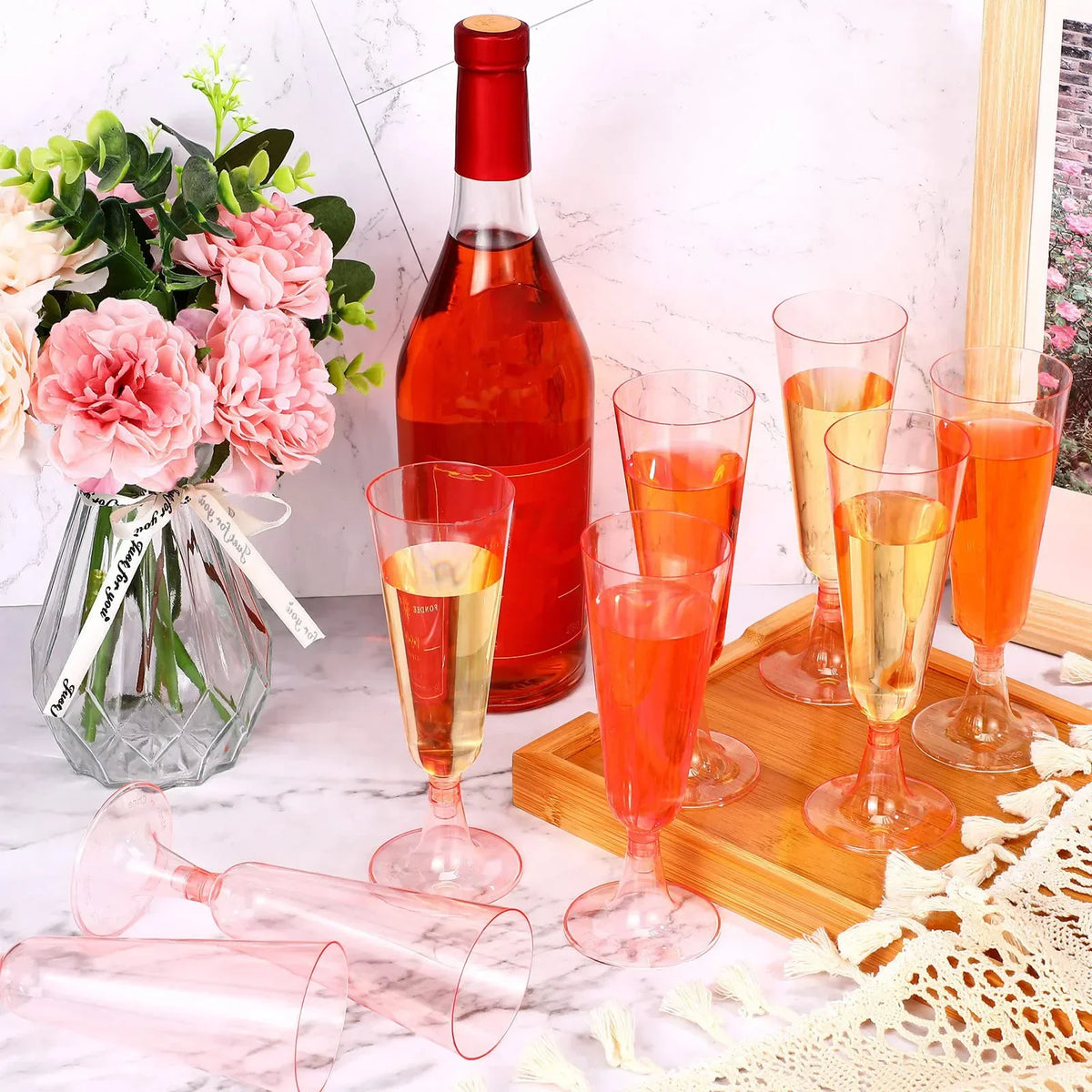 A set of disposable champagne glasses with a gold finish are displayed on a wooden tray next to pink flowers and other glassw