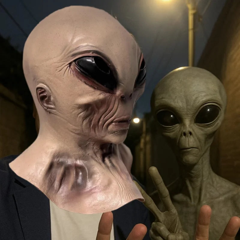 A realistic cute alien full-face latex mask with large black eyes and pale skin is shown against a dark background.